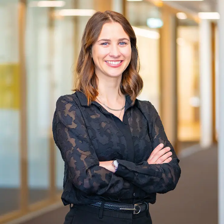 Junge Frau mit dunkler Bluse steht lächelnd mit verschränkten Armen im Büroflur von Sutter Medizintechnik – Team Einkauf.