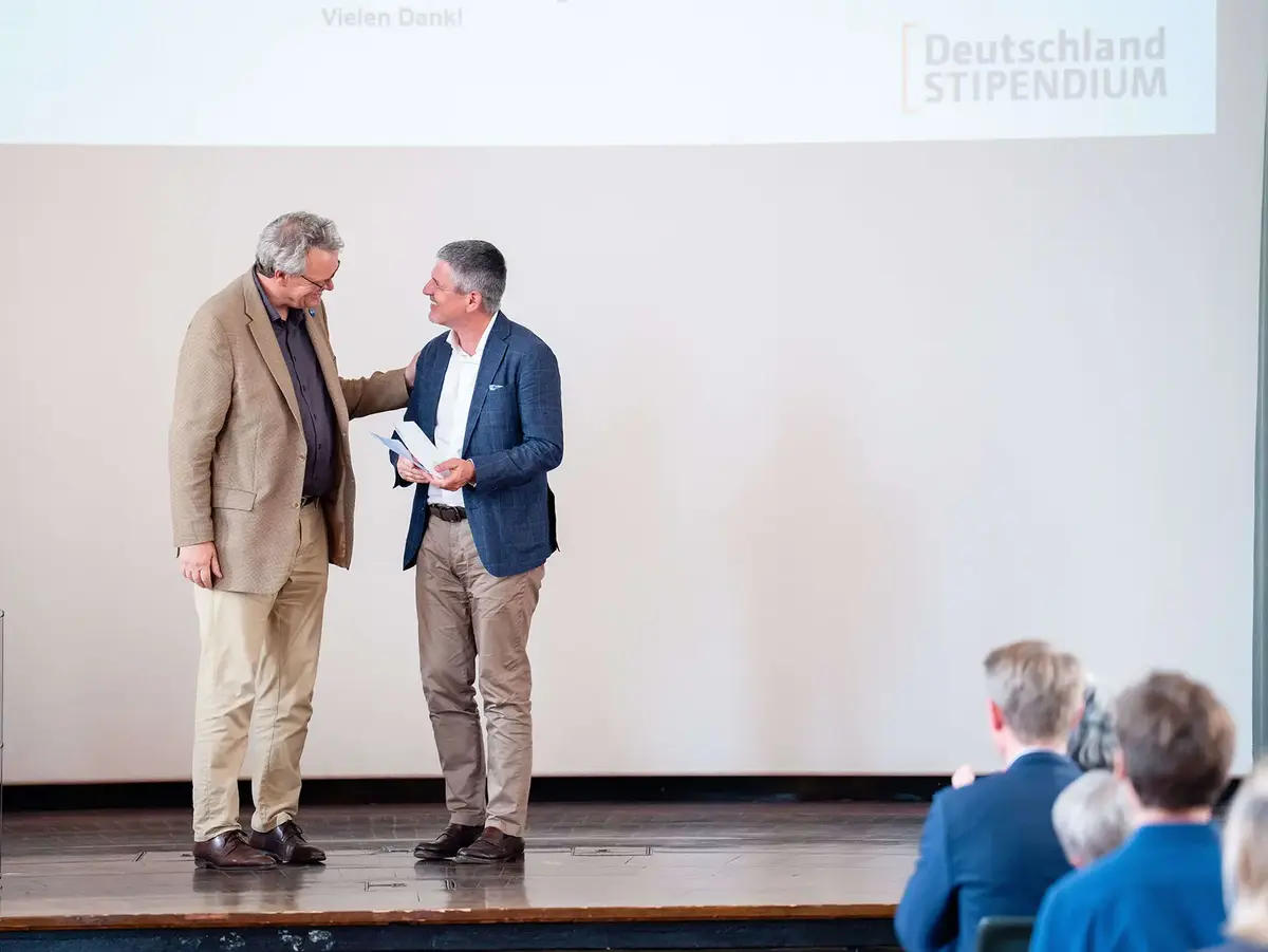 Feierliche Ehrung im Rahmen eines Stipendien- oder Förderprogramms auf einer Veranstaltungsbühne. Die Aufnahme zeigt die Übergabe von Dokumenten während einer offiziellen Zeremonie und steht für Anerkennung, Bildung und Engagement.