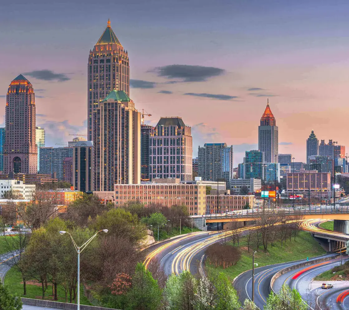 Skyline von Atlanta, USA, bei Sonnenuntergang mit beleuchteten Hochhäusern und fließendem Abendverkehr auf den geschwungenen Highways im Vordergrund.