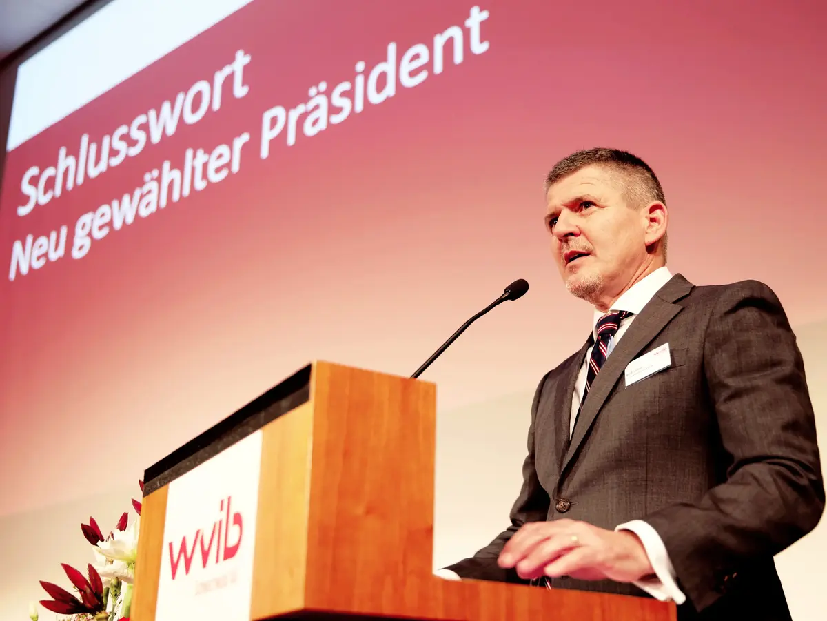 Person spricht an einem Rednerpult vor einer Veranstaltungsbühne mit rotem Hintergrund.