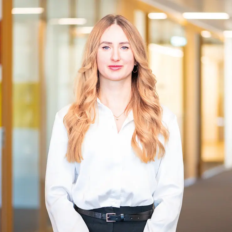 Junge Frau mit langen, blonden Haaren, weißer Bluse und dunkler Hose steht im Büroflur der Sutter Medizintechnik GmbH und blickt freundlich in die Kamera.