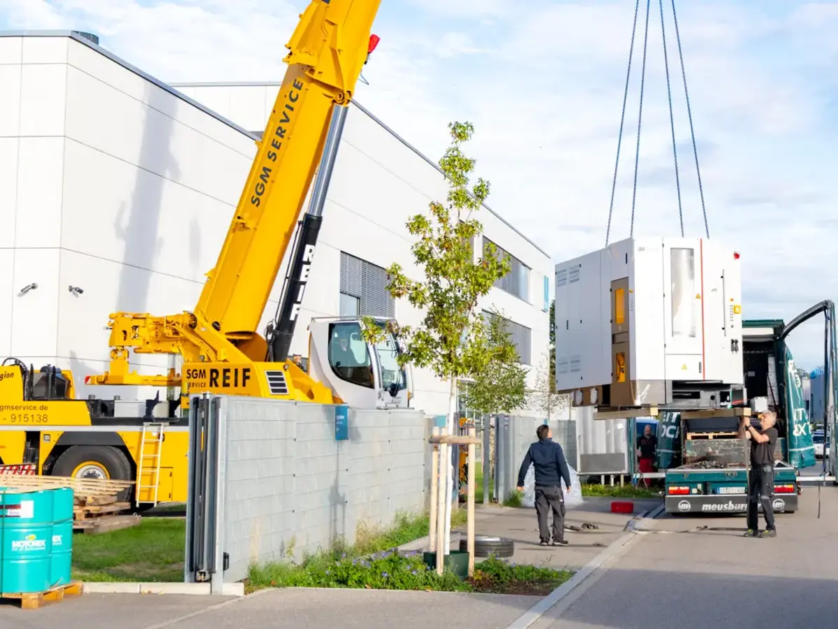Mobilkran hebt CNC-Produktionsanlage aus Lkw-Anhänger vor Industriegebäude.