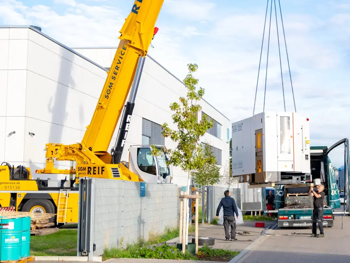 Mobile crane lifting CNC production equipment from a transport trailer at an industrial site.