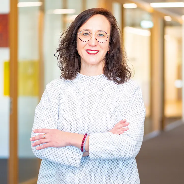 Customer Service Ansprechpartnerin steht mit verschränkten Armen im Flur eines modernen Bürogebäudes und lächelt in die Kamera.