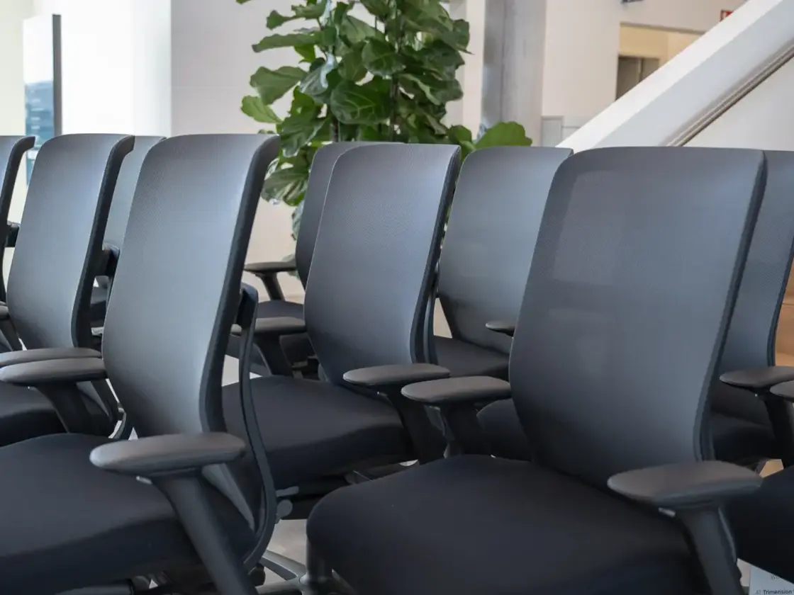 Reihe schwarzer Bürostühle mit Netzrücken und Armlehnen in einem Büro oder Konferenzraum, im Hintergrund große grüne Pflanze und Treppe