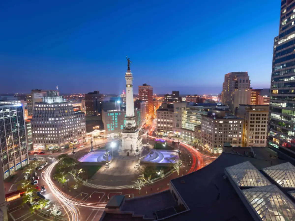 Aerial view of a city at dusk featuring an illuminated monument, surrounding buildings, and light trails from moving vehicles