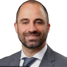 Porträtaufnahme einer Person mit kurzem Haar und Bart in grauem Sakko, weißem Hemd und Krawatte vor weißem Hintergrund