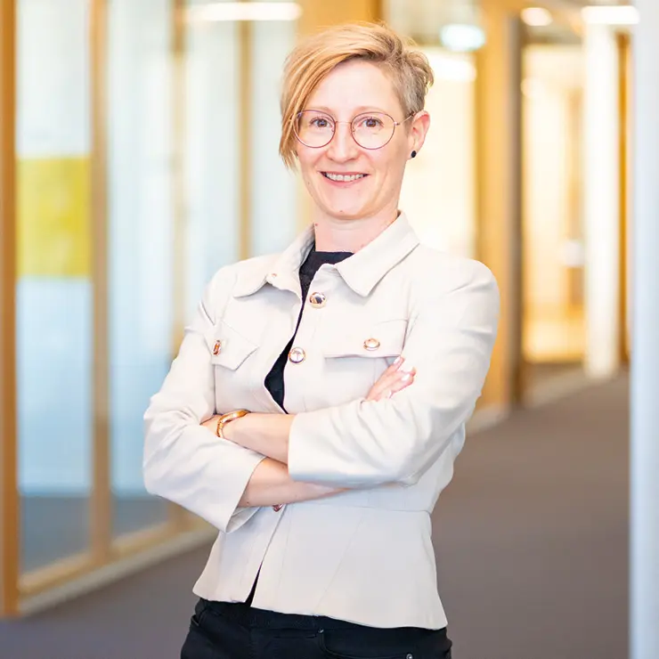 Frau mit kurzem, blondem Haar, heller Jacke und Brille steht mit verschränkten Armen im Büroflur von Sutter Medizintechnik und lächelt in die Kamera.