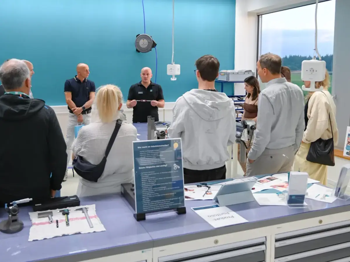Visitor group touring a training workshop with technical workstations and manufacturing equipment.