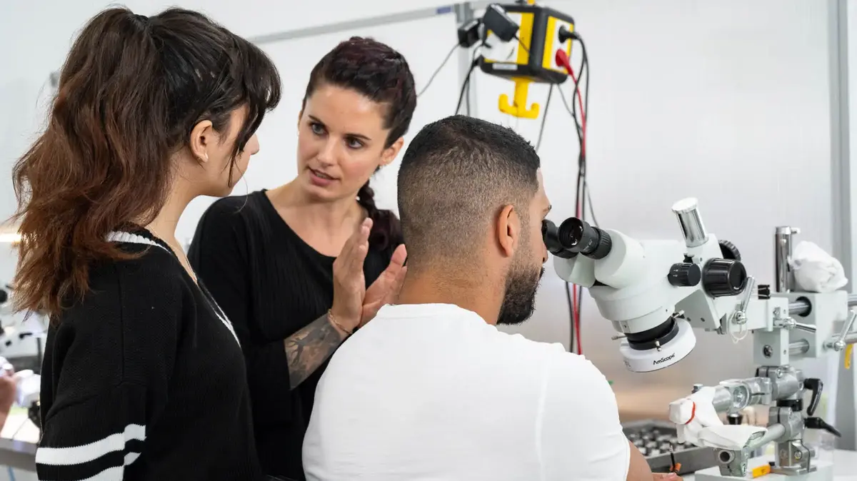 Ausbilderin erklärt Auszubildenden bei Sutter Medizintechnik die Arbeit an einem Mikroskoparbeitsplatz – praxisnahe Ausbildung in der Feinmechanik.