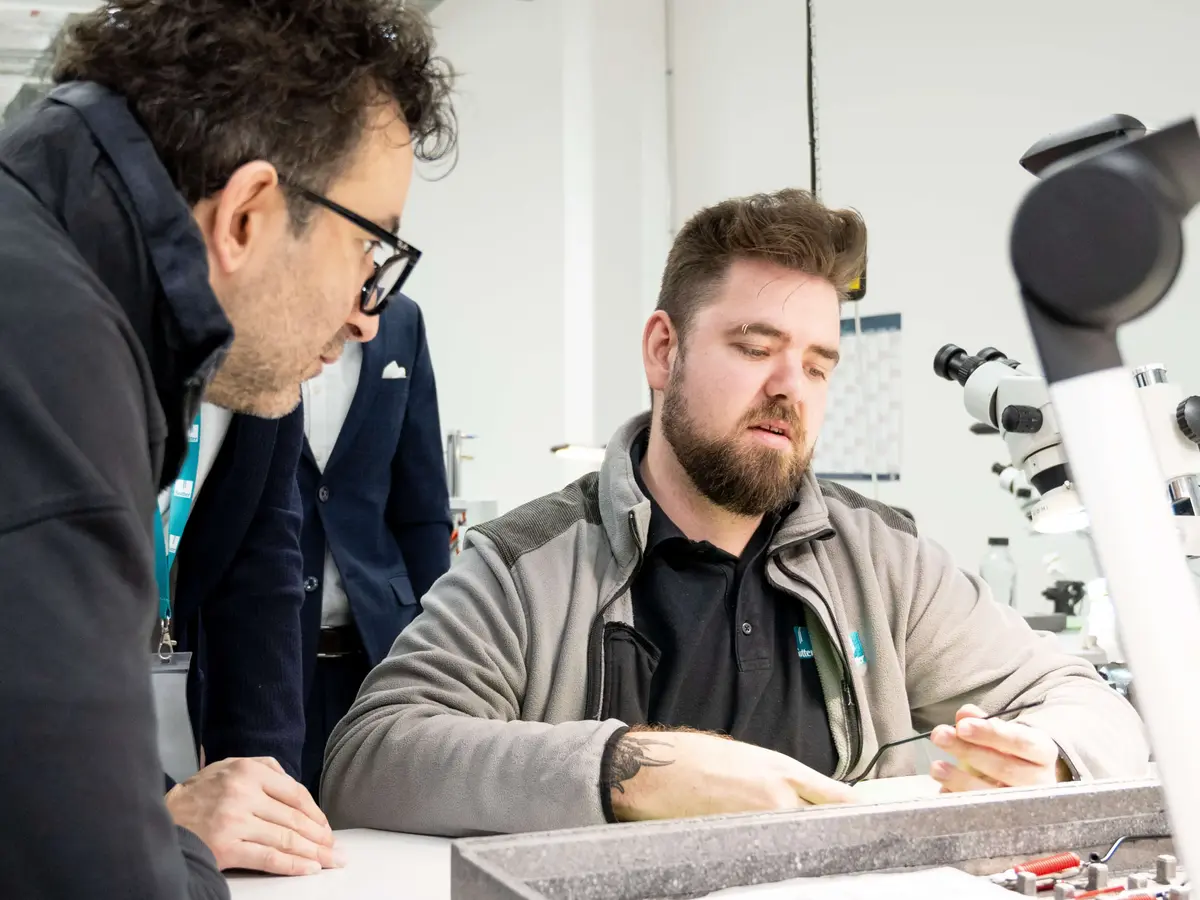 Zwei Mitarbeitende prüfen gemeinsam ein Bauteil an einem Arbeitsplatz mit Mikroskop in einem technischen Umfeld.