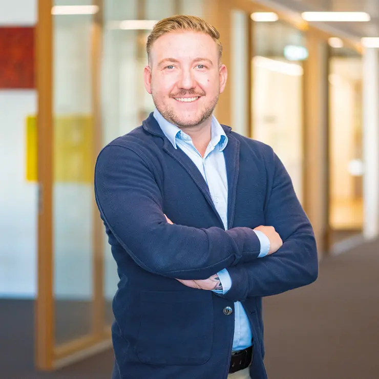 Ein lächelnder Mitarbeiter von Sutter Medizintechnik steht mit verschränkten Armen im Büroflur und trägt ein dunkelblaues Sakko über einem hellblauen Hemd.