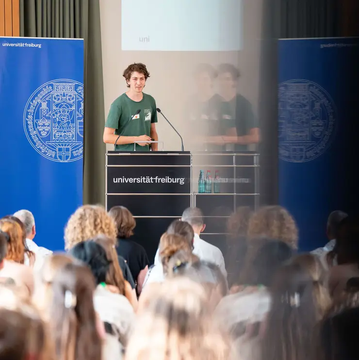Speaker presenting at a University of Freiburg event in front of a large audience. The image highlights academic exchange, student engagement, and active participation in scholarship and support programs.