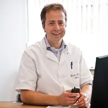 Dr. Geukens in weißem Arztkittel an einem Schreibtisch, lächelnd in die Kamera.