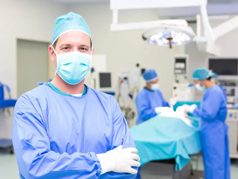 Male surgeon in surgical attire and face mask stands in the foreground, while two colleagues operate at the OR table in the background.