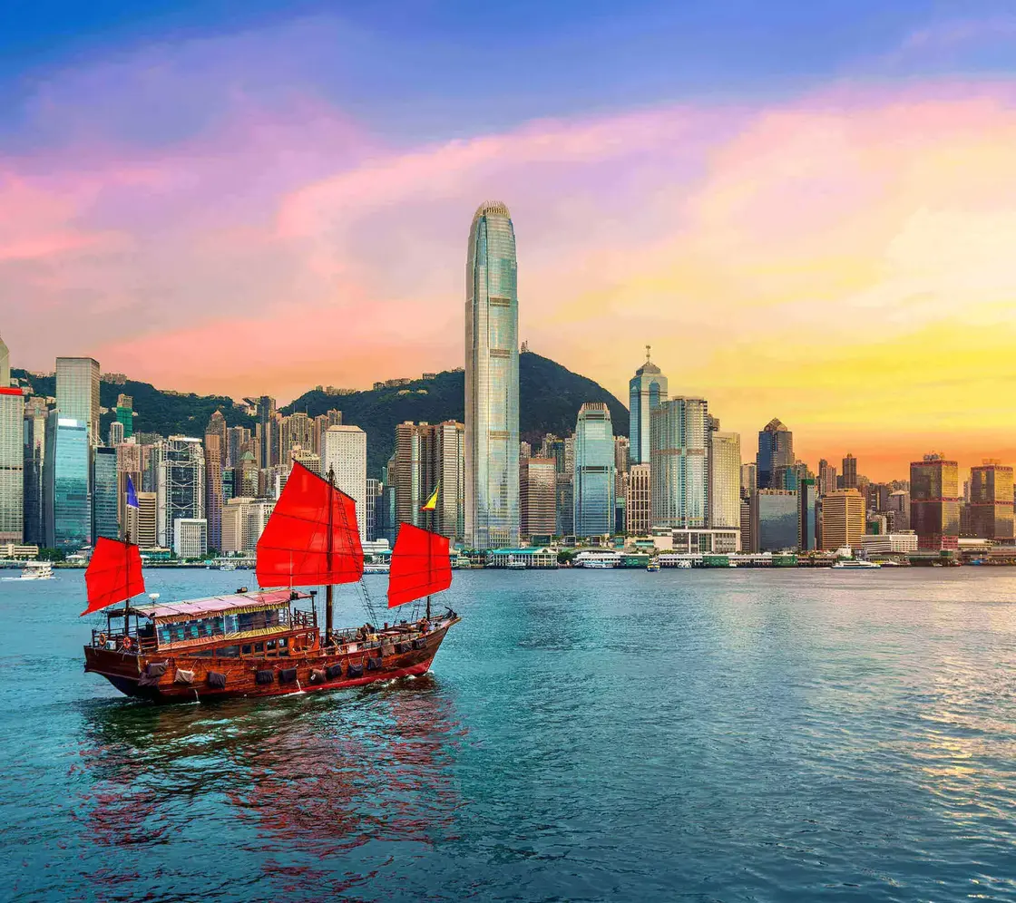 Traditionelles chinesisches Junkboot mit roten Segeln fährt vor der Skyline von Hongkong bei farbenprächtigem Sonnenuntergang über das Wasser.        