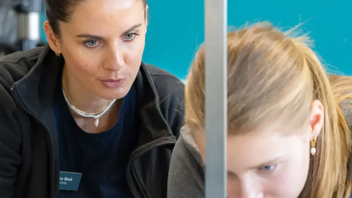 Junge Teilnehmerin des Girls’Day misst mit Unterstützung einer Sutter-Mitarbeiterin ein Werkstück in der Fertigung; beide konzentriert bei der Arbeit vor türkisfarbener Wand.