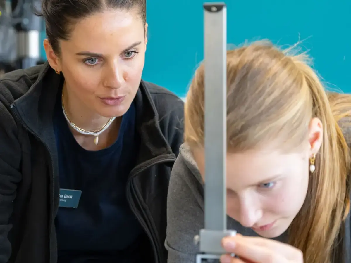Junge Teilnehmerin des Girls’Day misst mit Unterstützung einer Sutter-Mitarbeiterin ein Werkstück in der Fertigung; beide konzentriert bei der Arbeit vor türkisfarbener Wand.