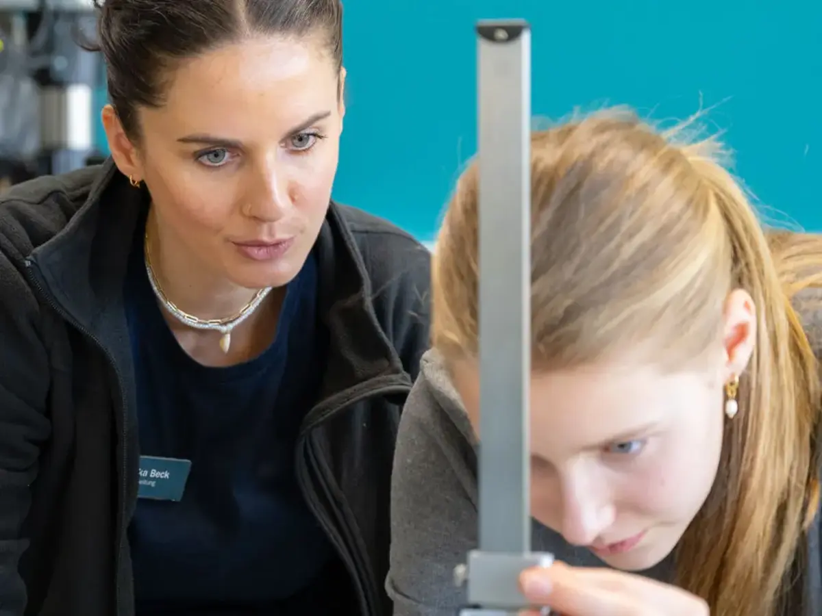 Junge Teilnehmerin des Girls’Day misst mit Unterstützung einer Sutter-Mitarbeiterin ein Werkstück in der Fertigung; beide konzentriert bei der Arbeit vor türkisfarbener Wand.