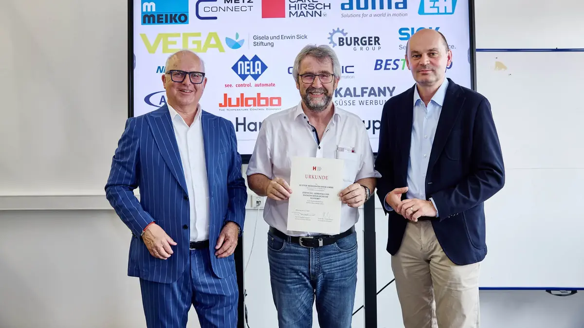 Drei Männer bei einer Urkundenübergabe vor einer Sponsorenwand am Robotiktag 2025; ein Vertreter von Sutter Medizintechnik hält die Urkunde der Hochschule Offenburg.