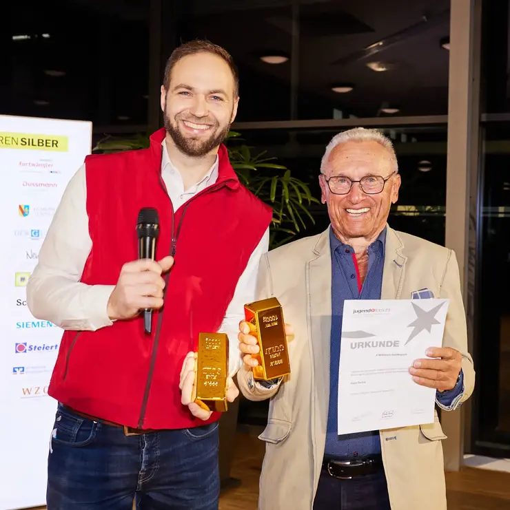 Zwei Personen mit Urkunde und symbolischem Preis bei einer Auszeichnung auf einer Veranstaltung.