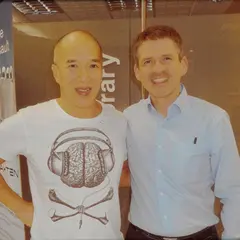 Gerahmtes Foto von zwei Personen, die nebeneinander stehen. Eine Person trägt einen weißen Hoodie mit Grafikdesign, die andere ein hellblaues Hemd. Beide lächeln und befinden sich in einem Innenraum mit Dekoration im Hintergrund.