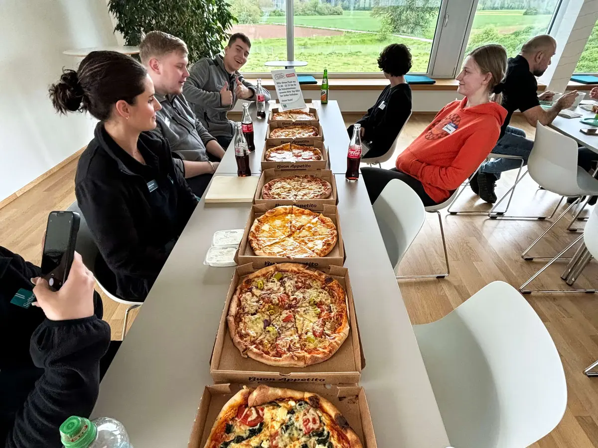 Teilnehmende und Mitarbeitende sitzen bei einem gemeinsamen Pizza-Mittagessen in einem modernen Aufenthaltsraum.