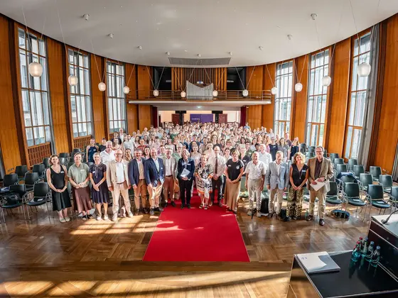 Large group photo taken during an event in a bright conference hall featuring tall windows, wood-paneled walls, and a red carpet. The image conveys collaboration, community, and a professional event setting.
