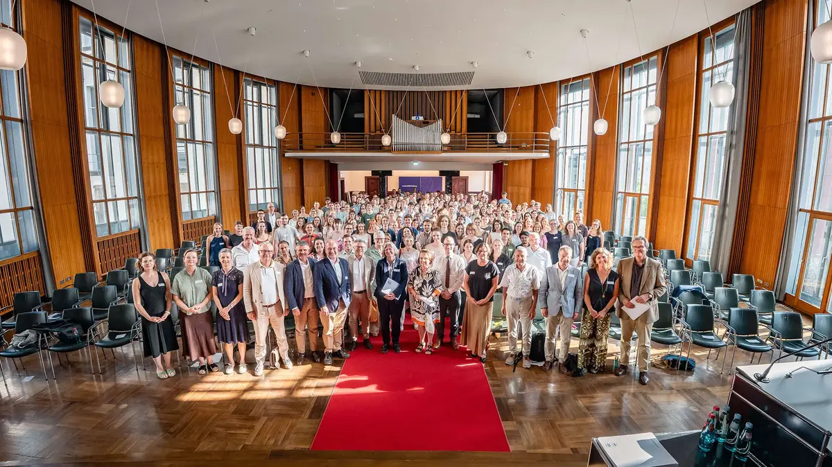 Large group photo taken during an event in a bright conference hall featuring tall windows, wood-paneled walls, and a red carpet. The image conveys collaboration, community, and a professional event setting.