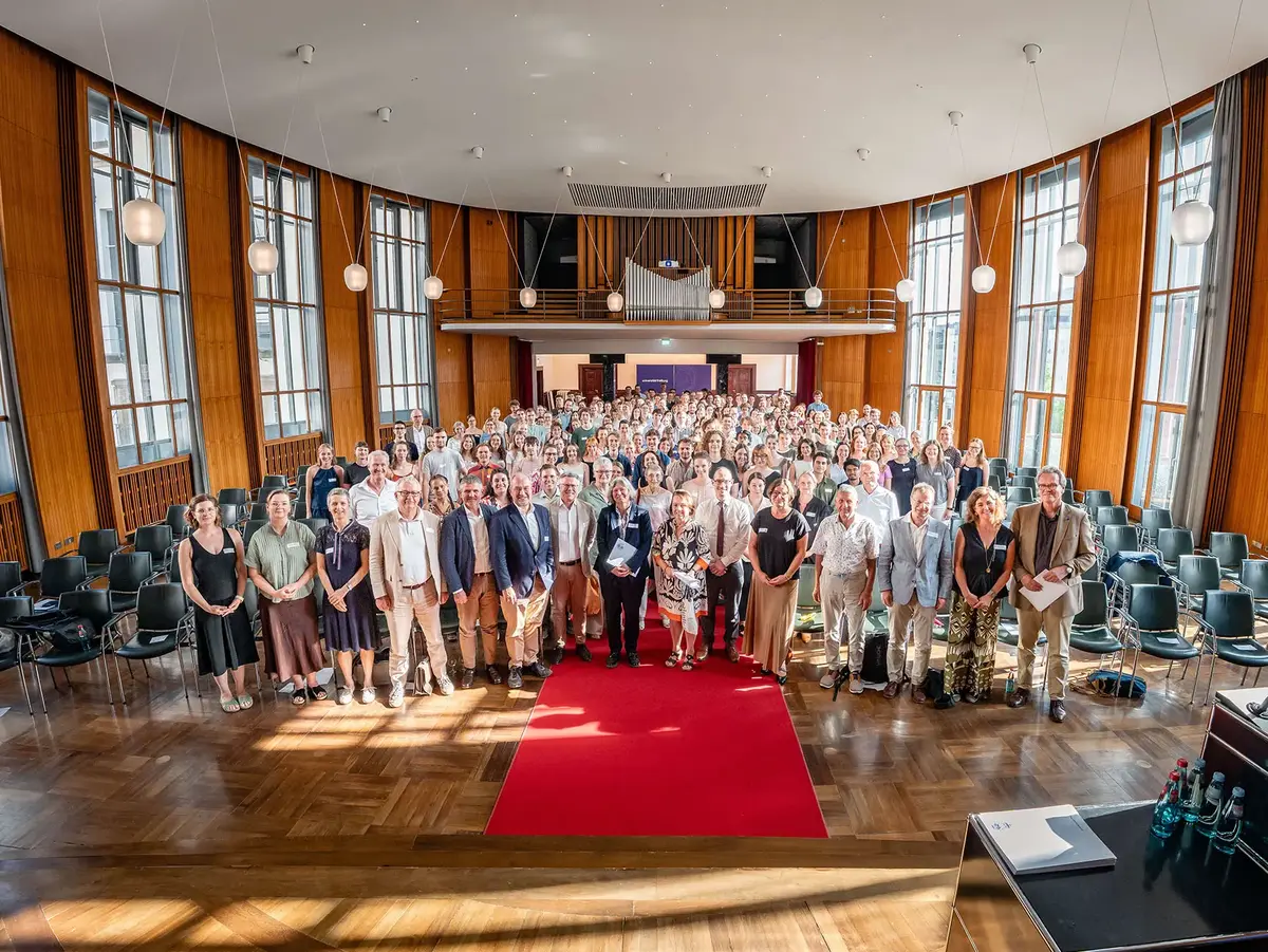Großes Gruppenfoto einer Veranstaltung in einem lichtdurchfluteten Saal mit hohen Fenstern, Holzvertäfelung und rotem Teppich. Die Aufnahme vermittelt Gemeinschaft, Austausch und eine professionelle Veranstaltungsatmosphäre.