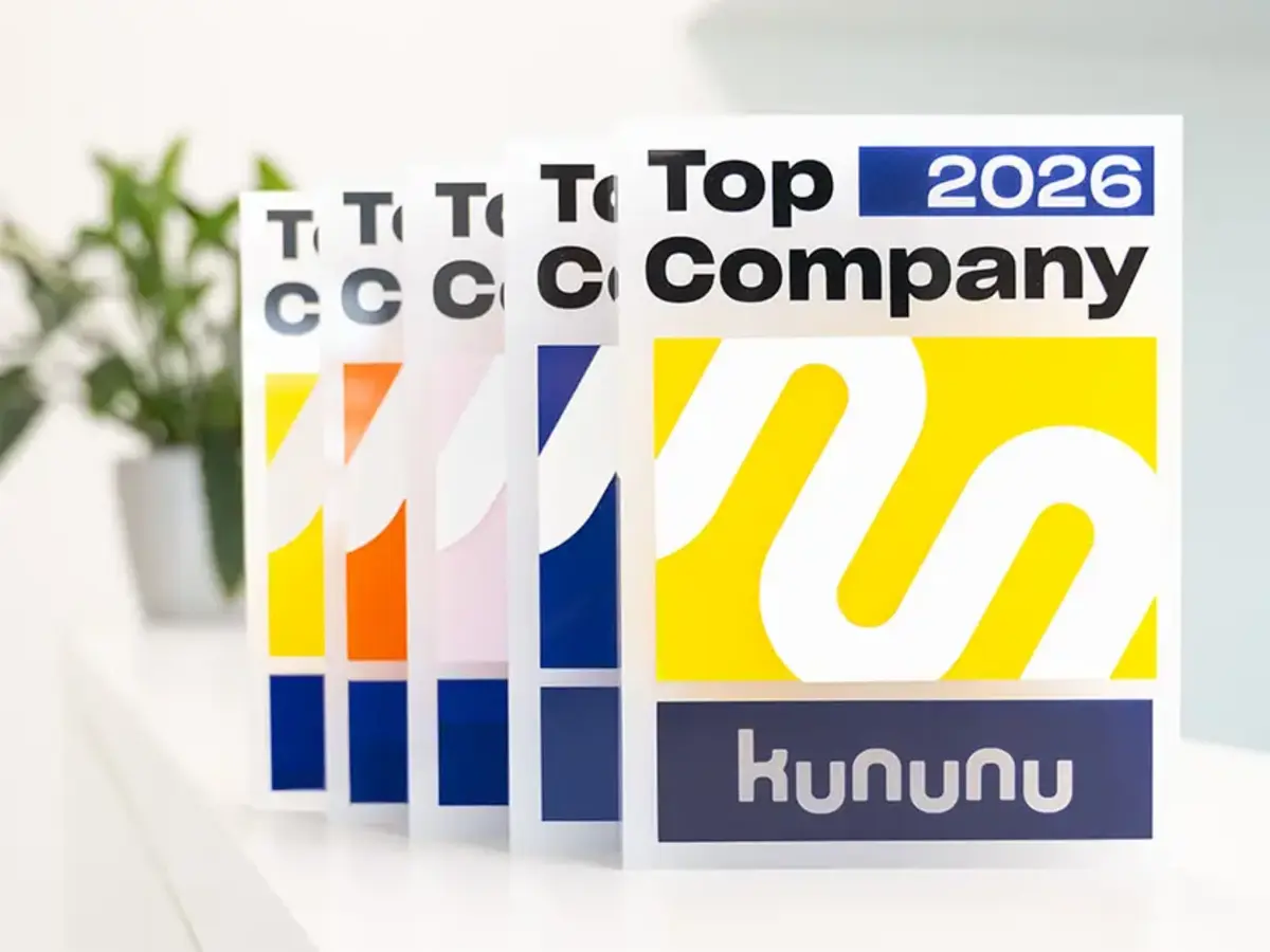 Mehrere kununu‑„Top Company 2026“-Auszeichnungen stehen nebeneinander auf einer weißen Fläche. Jede Auszeichnung zeigt ein gelbes und blaues grafisches Motiv sowie den Schriftzug „Top Company 2026“ und das kununu‑Logo. Im Hintergrund befindet sich ein heller, moderner Bürobereich mit einer Pflanze