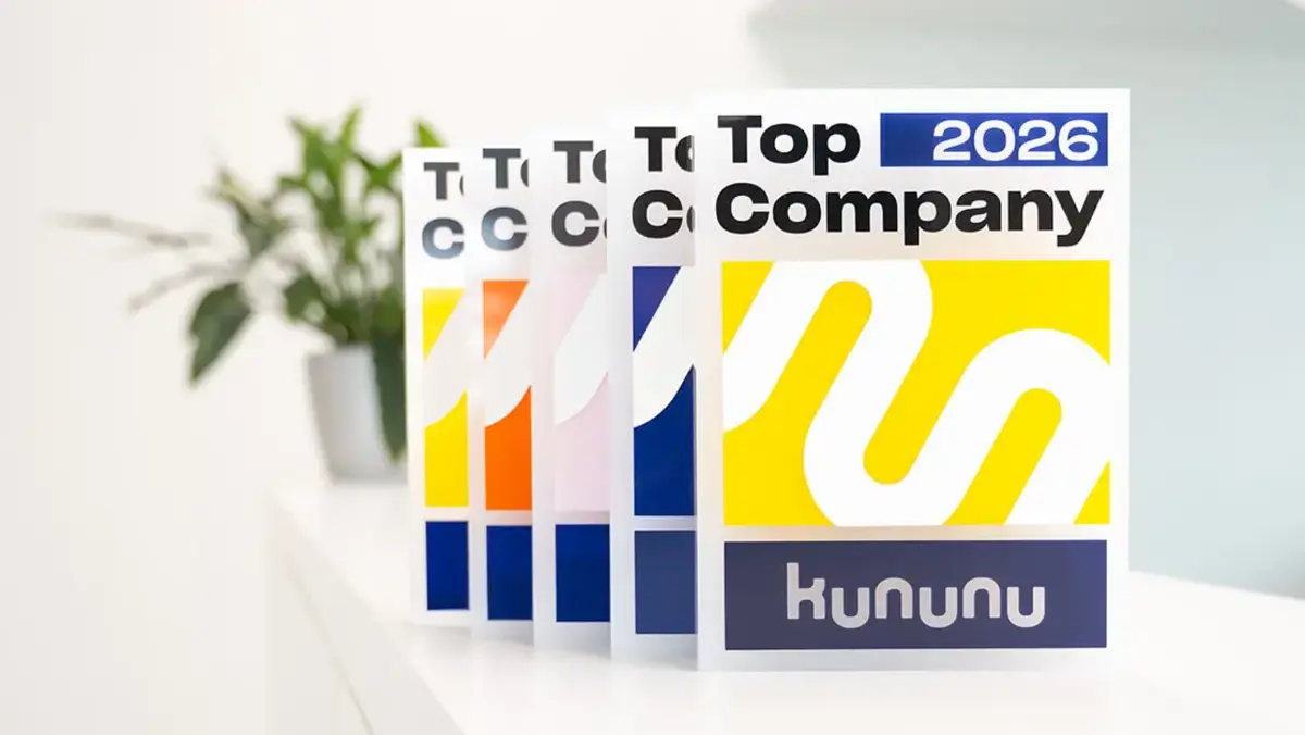 Multiple kununu “Top Company 2026” awards displayed in a row on a white surface, featuring a yellow‑and‑blue graphic design, the “Top Company 2026” label, and the kununu logo. A bright, modern office environment with a plant appears in the background.
