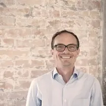 Person in hellblauem Hemd lächelnd vor einer Backsteinwand