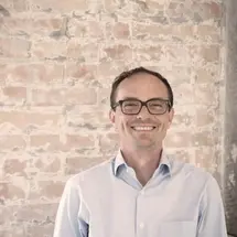 Person in hellblauem Hemd lächelnd vor einer Backsteinwand