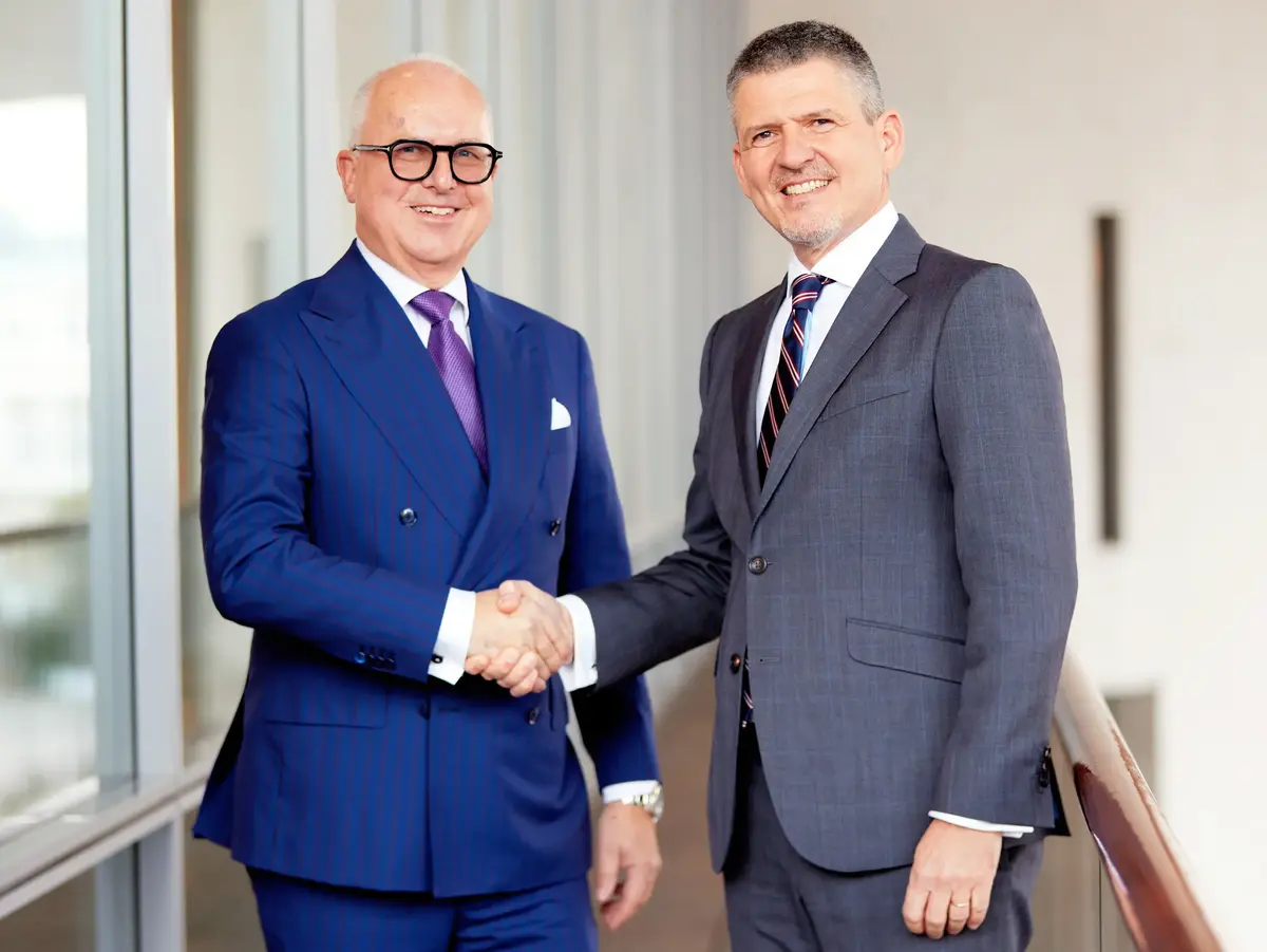 Two business professionals in suits shaking hands inside a modern office building.