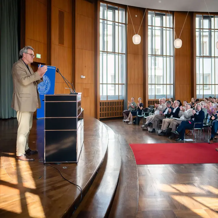 Ceremonial event of the Deutschlandstipendium at the University of Freiburg held in a bright auditorium. The image highlights a keynote speech in front of a large audience and reflects educational support, academic engagement, and talent development.