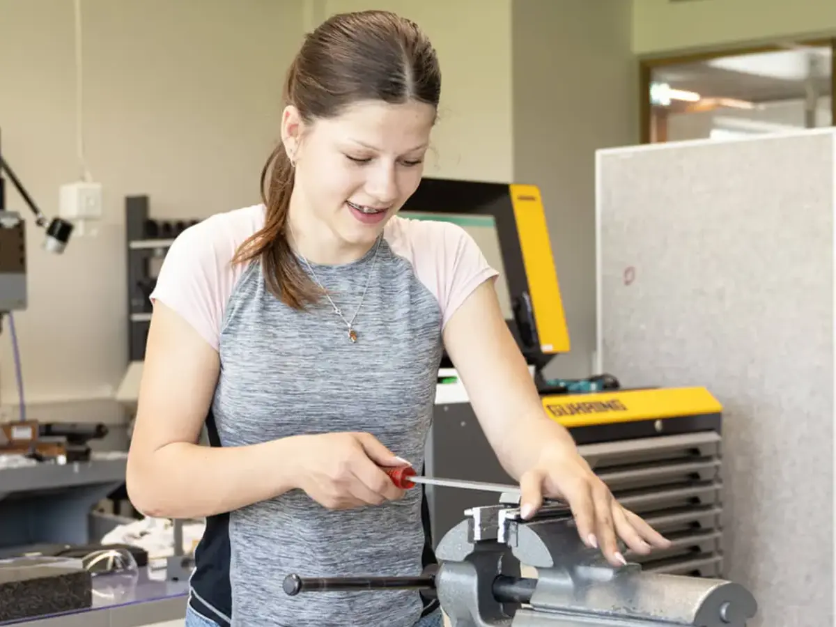 Praktische technische Ausbildung in einer modernen Werkstatt: Person bearbeitet ein Werkstück mit Schraubendreher und Schraubstock an einer Werkbank.