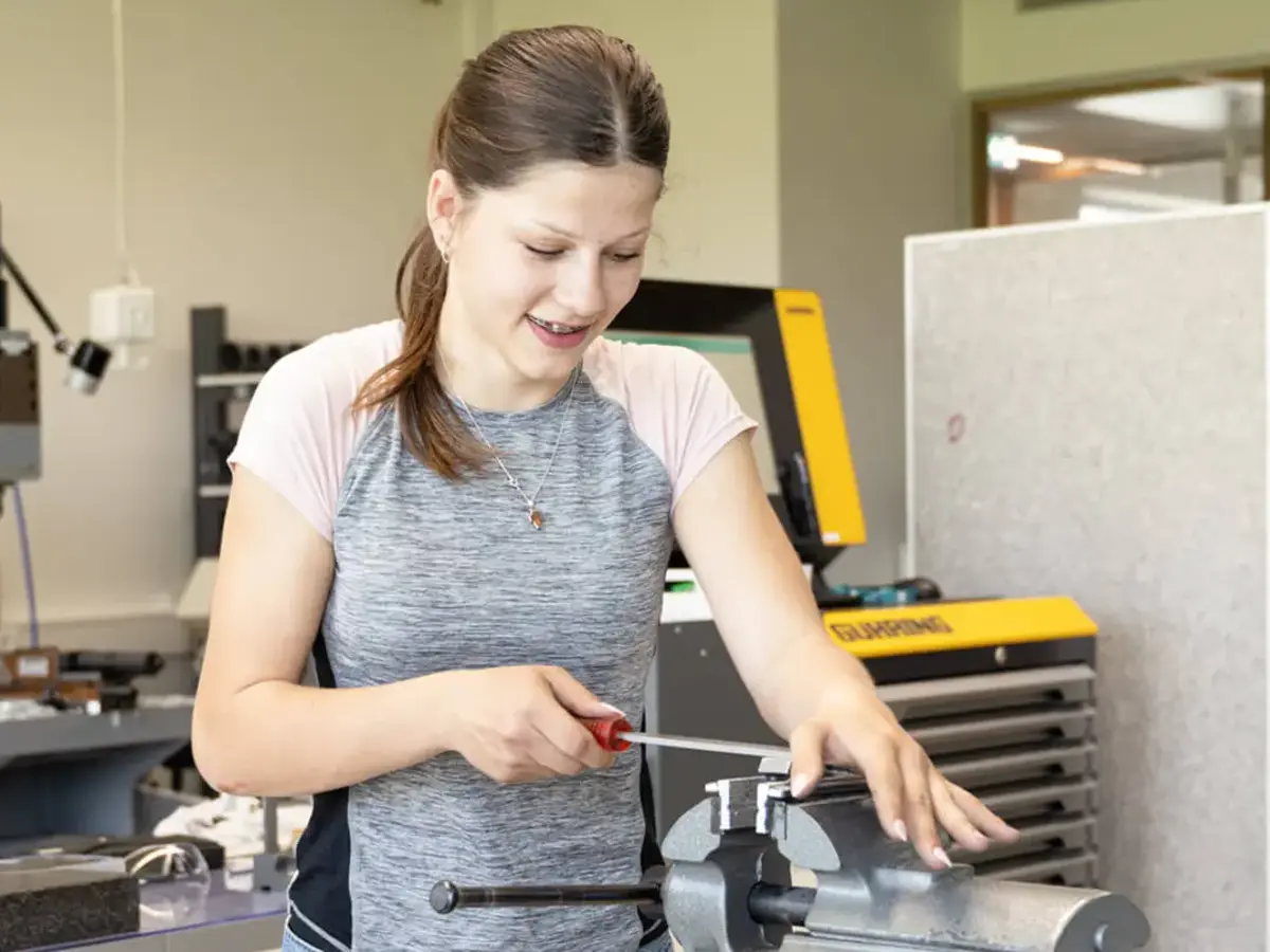 Hands-on technical training in a modern workshop: person working on a clamped component using a screwdriver and bench vise.