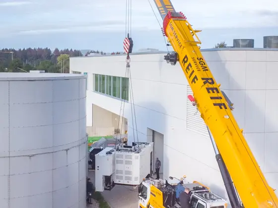 Mobile crane positioning industrial machinery between production buildings at a manufacturing site.