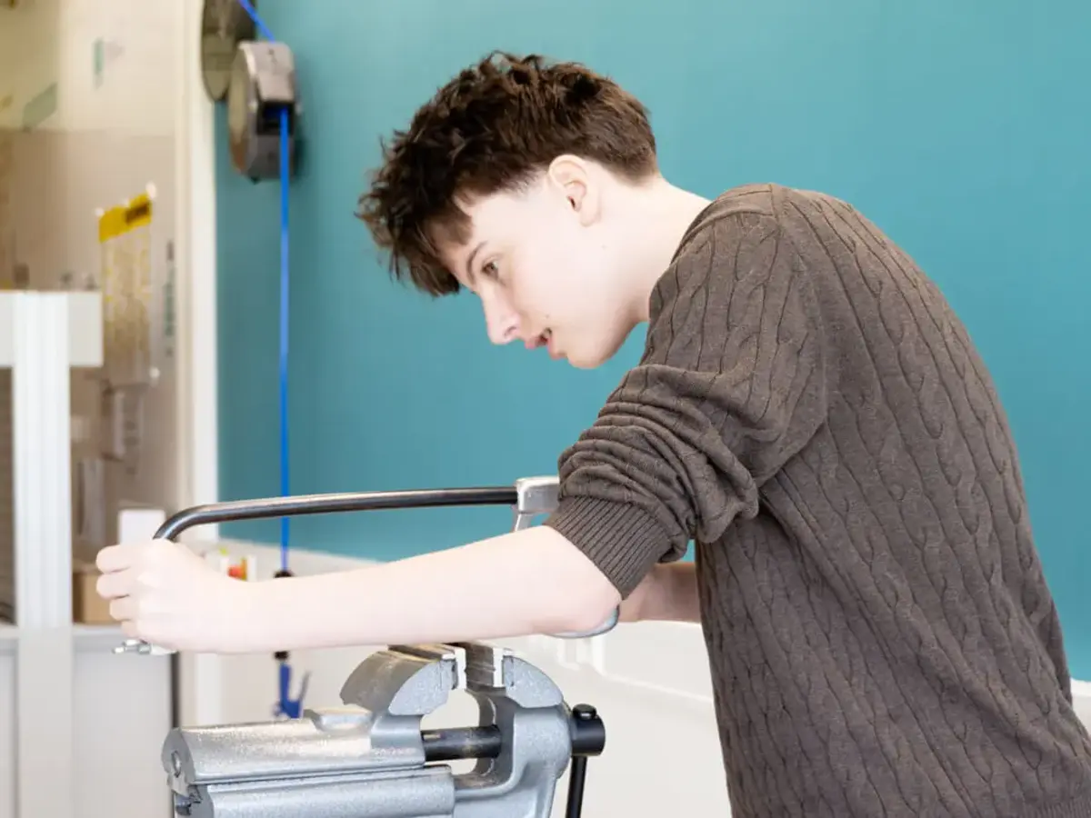 Person performing hands-on metalworking training in a workshop using a hand saw and bench vise at a workbench.