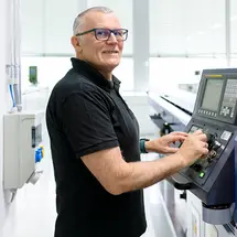 Ein Mitarbeiter von Sutter Medizintechnik bedient eine CNC-Drehmaschine in der Fertigung; er trägt ein schwarzes Poloshirt und steht vor einem Bedienfeld.