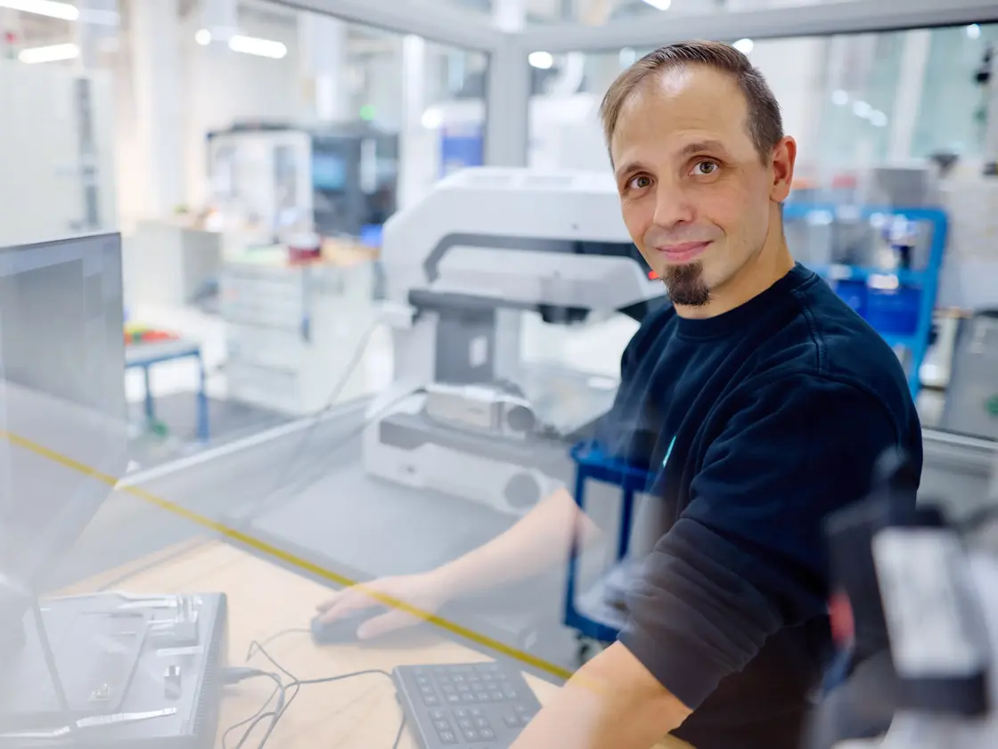 Sutter_Medizintechnik_2025_Keyence_CNC © Jörg Wilhelm Mitarbeiter von Sutter Medizintechnik steht an einem Arbeitsplatz in der CNC und blickt lächelnd in die Kamera. Im Hintergrund sind moderne Prüfgeräte und Fertigungsbereiche zu sehen.