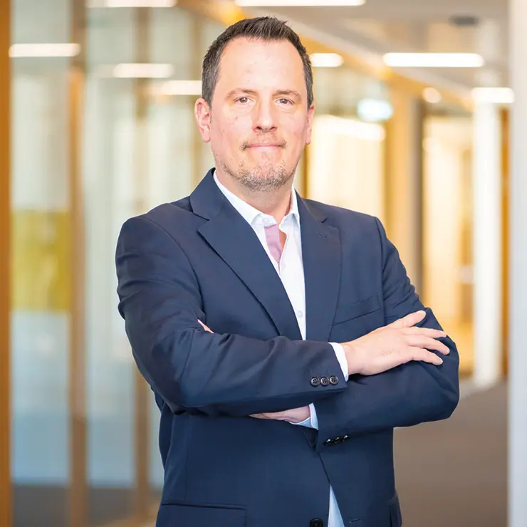 Ein Mann im dunkelblauen Anzug mit rosa Krawatte steht mit verschränkten Armen im Flur der Sutter Medizintechnik GmbH und blickt ernst in die Kamera.