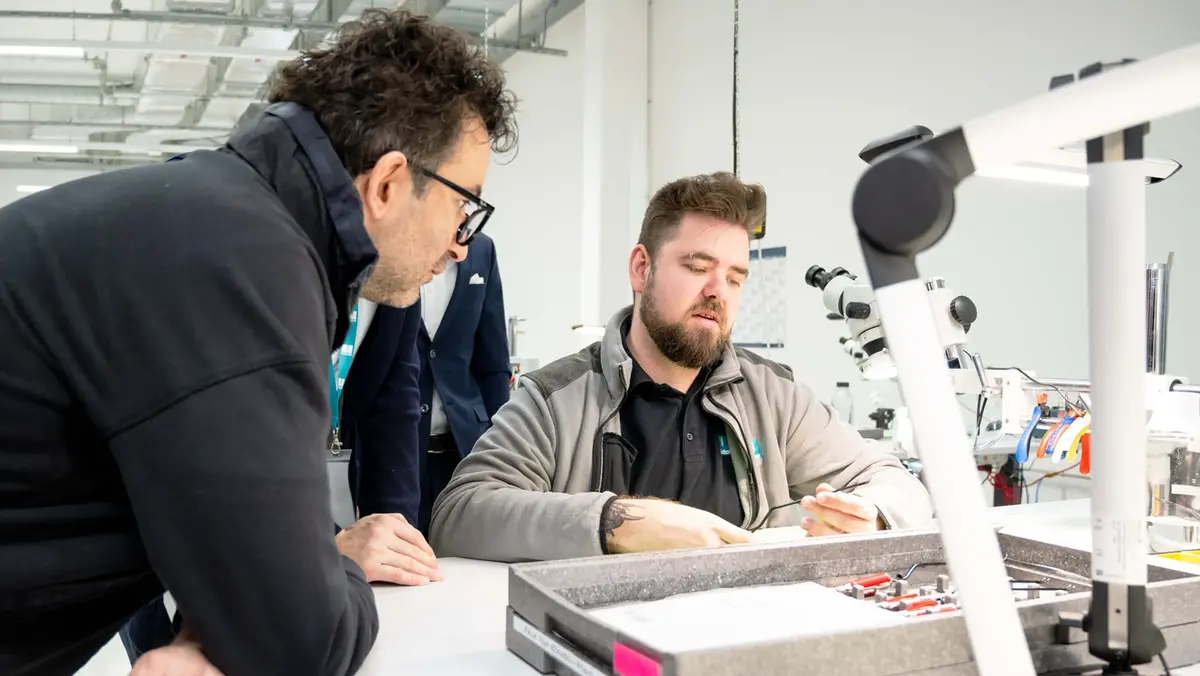 Personen im Gespräch an einem Werkstatttisch mit feinmechanischen Instrumenten und Arbeitsplätzen im Hintergrund.