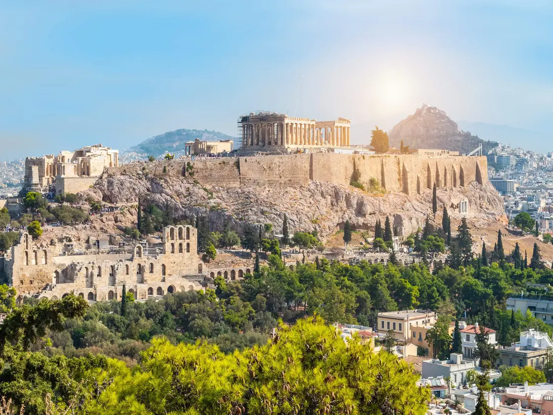 Blick auf die Akropolis in Athen mit dem Parthenon-Tempel und umliegenden antiken Bauwerken – im Hintergrund moderne Stadtlandschaft und grüne Hügel.