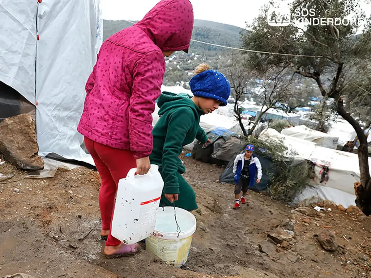 Zwei Kinder im Moria Camp tragen barfuß und in warmer Kleidung Kanister und Eimer mit Wasser einen matschigen Hang hinauf; im Hintergrund weitere Kinder und notdürftige Zelte, Logo von SOS-Kinderdorf.